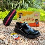 Girls' black patent buckle school shoes shown on gravel beside the shoe box