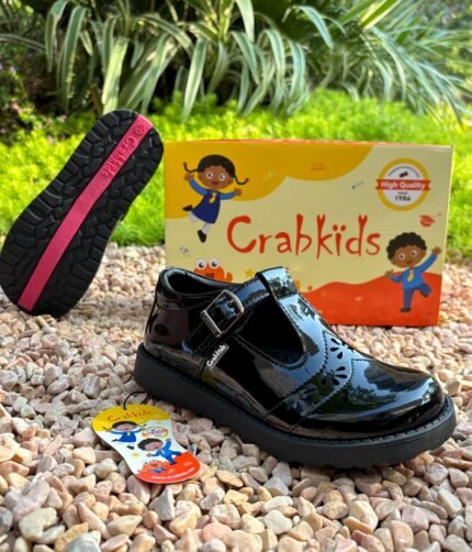 Girls' black patent buckle school shoes shown on gravel beside the shoe box